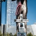 A memorial tree at the 9/11 Memorial in New York City, decorated with tributes marking the 25th anniversary of the September 11 attacks. The tree bears a 'Never 25 Forget' sign, a memorial poster for P.O. Paul Talty of the NYPD Emergency Service Unit, and a group tribute sheet titled 'Our NYPD Heroes' featuring photos of fallen officers. Artificial flowers, an American flag, and a '911' placard are also attached to the tree.