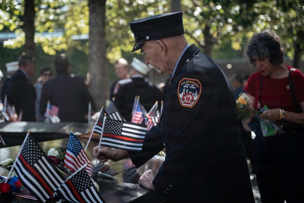 A Fire Department City of New York (FDNY) firefighter in dress uniform bows his head at what appears to be the 9/11 Memorial in New York City. The memorial's bronze parapet is lined with small American flags, including Thin Red Line flags symbolizing fallen firefighters, along with flowers. The scene depicts a solemn commemorative ceremony with multiple uniformed personnel and civilians present.