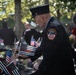 A Fire Department City of New York (FDNY) firefighter in dress uniform bows his head at what appears to be the 9/11 Memorial in New York City. The memorial's bronze parapet is lined with small American flags, including Thin Red Line flags symbolizing fallen firefighters, along with flowers. The scene depicts a solemn commemorative ceremony with multiple uniformed personnel and civilians present.