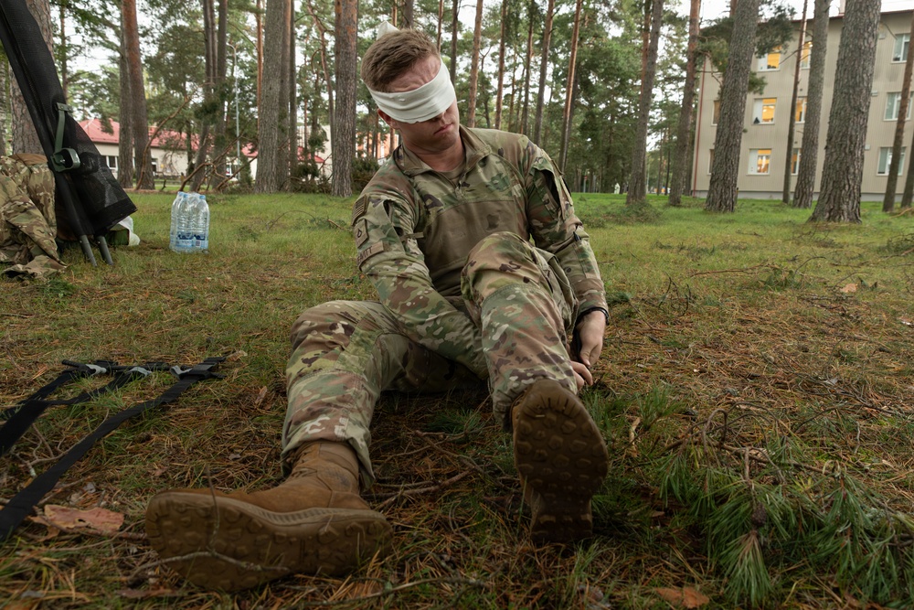 A soldier in US Army MultiCam (OCP) camouflage uniform is seated on the ground in a wooded area with a white blindfold covering his eyes. His hands appear to be restrained behind his back, suggesting this is a SERE (Survival, Evasion, Resistance, and Escape) training exercise or prisoner-of-war simulation. Buildings are visible in the background, indicating a military installation or training base.