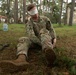 A soldier in US Army MultiCam (OCP) camouflage uniform is seated on the ground in a wooded area with a white blindfold covering his eyes. His hands appear to be restrained behind his back, suggesting this is a SERE (Survival, Evasion, Resistance, and Escape) training exercise or prisoner-of-war simulation. Buildings are visible in the background, indicating a military installation or training base.