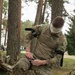 A soldier in MultiCam/OCP camouflage uniform is kneeling on grass in a wooded area, applying or adjusting a tourniquet on his own leg. He has a white bandage wrapped around his head/eyes, suggesting a simulated casualty training exercise. The setting appears to be a military training environment with trees and a road visible in the background.