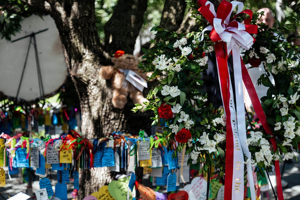 The image depicts an outdoor memorial or tribute site centered around a large tree. A formal floral wreath with red and white ribbons is prominently displayed on the right, while the tree and surrounding fence are covered with colorful handwritten notes, tags, and ribbons. A stuffed teddy bear is also visible tied to the tree, suggesting this is a public grief or remembrance site.