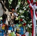 The image depicts an outdoor memorial or tribute site centered around a large tree. A formal floral wreath with red and white ribbons is prominently displayed on the right, while the tree and surrounding fence are covered with colorful handwritten notes, tags, and ribbons. A stuffed teddy bear is also visible tied to the tree, suggesting this is a public grief or remembrance site.