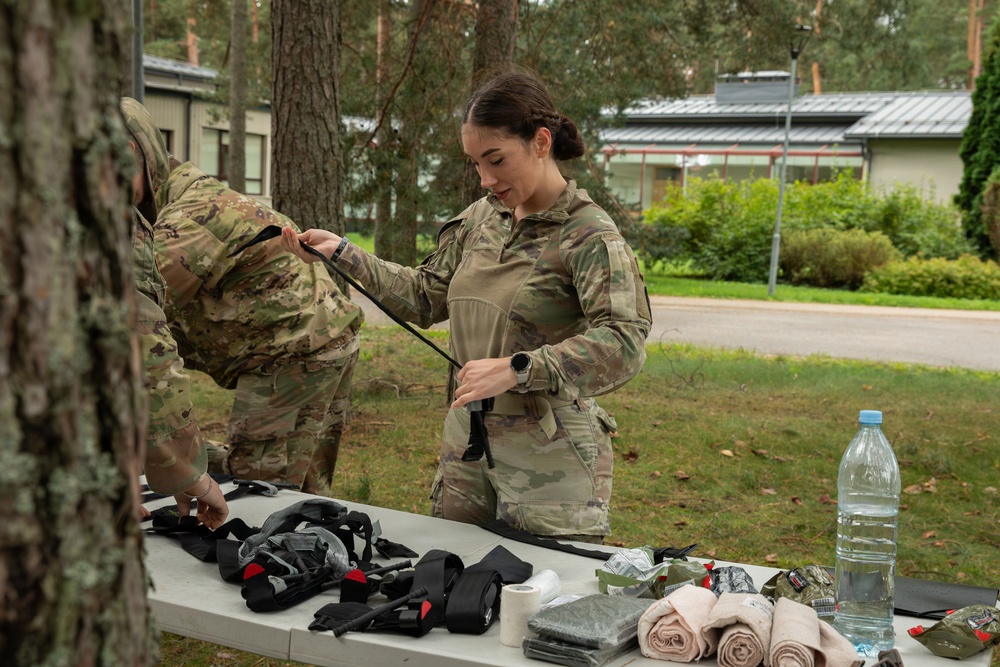 Two military personnel in OCP (Operational Camouflage Pattern) uniforms are working at an outdoor table, handling tactical equipment. A female soldier in the foreground appears to be adjusting or demonstrating a strap or harness, while a second soldier assists. The setting appears to be a military training area or base, with trees and a building visible in the background.