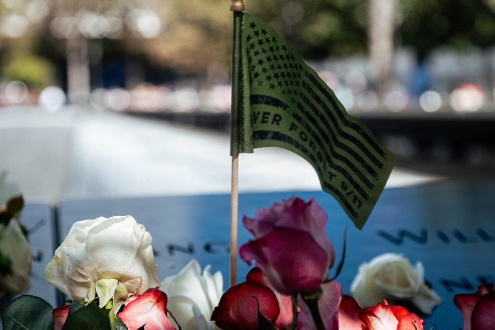 A close-up photograph of a 9/11 memorial site showing flowers and a commemorative flag placed on an engraved memorial panel. A small American-style flag bearing the text 'NEVER FORGET 9/11' is prominently displayed among white, red, and pink roses. The background shows a blurred reflective pool, consistent with the 9/11 Memorial in New York City.