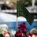 A close-up photograph of a 9/11 memorial site showing flowers and a commemorative flag placed on an engraved memorial panel. A small American-style flag bearing the text 'NEVER FORGET 9/11' is prominently displayed among white, red, and pink roses. The background shows a blurred reflective pool, consistent with the 9/11 Memorial in New York City.