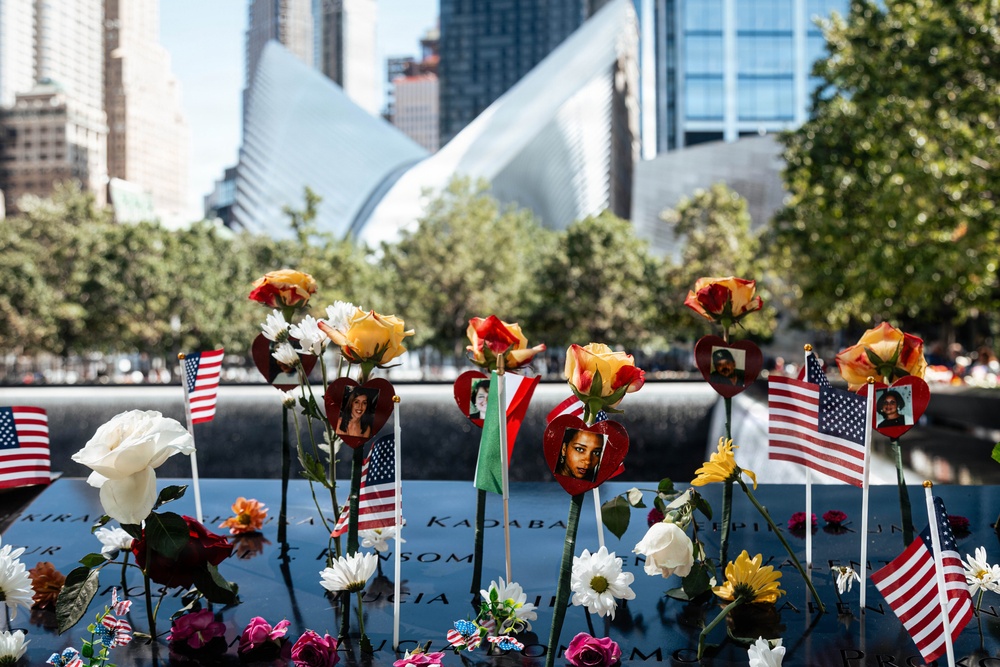 The image shows the 9/11 Memorial in Lower Manhattan, New York City, with flowers, American flags, and heart-shaped photo frames placed along the engraved bronze parapet surrounding one of the memorial pools. The tributes include roses, daisies, sunflowers, and small photographs of victims placed in decorative red heart frames. The Oculus (World Trade Center Transportation Hub) and surrounding skyscrapers are visible in the background.