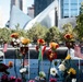 The image shows the 9/11 Memorial in Lower Manhattan, New York City, with flowers, American flags, and heart-shaped photo frames placed along the engraved bronze parapet surrounding one of the memorial pools. The tributes include roses, daisies, sunflowers, and small photographs of victims placed in decorative red heart frames. The Oculus (World Trade Center Transportation Hub) and surrounding skyscrapers are visible in the background.