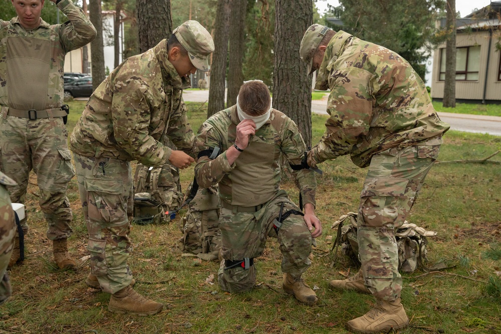 Military personnel in MultiCam/OCP camouflage uniforms are conducting what appears to be a combat casualty care or tactical first aid training exercise outdoors. Two soldiers are assisting a kneeling soldier who has a bandage applied to his face/head area, suggesting a simulated wound treatment scenario. The setting appears to be a military installation or training area with trees and buildings visible in the background.