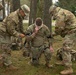 Military personnel in MultiCam/OCP camouflage uniforms are conducting what appears to be a combat casualty care or tactical first aid training exercise outdoors. Two soldiers are assisting a kneeling soldier who has a bandage applied to his face/head area, suggesting a simulated wound treatment scenario. The setting appears to be a military installation or training area with trees and buildings visible in the background.