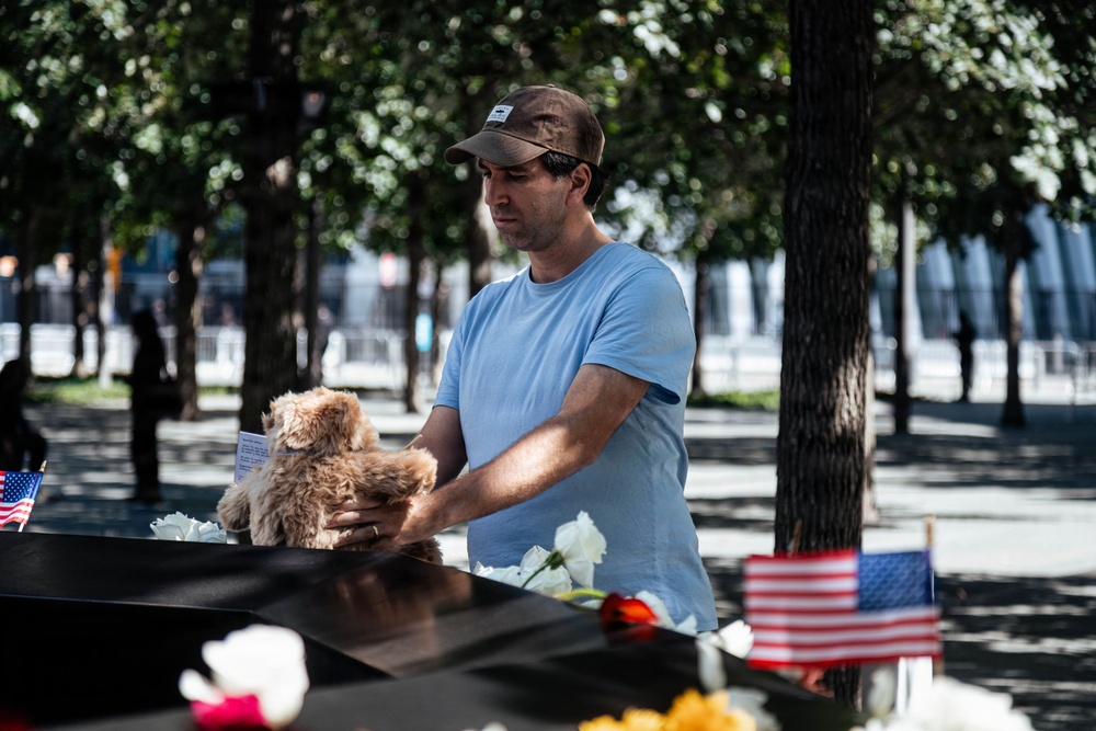 A man wearing a light blue t-shirt and brown baseball cap places a stuffed teddy bear on what appears to be the 9/11 Memorial in New York City. The dark granite memorial edge is adorned with flowers, small American flags, and other tributes. The man's downward gaze and deliberate gesture suggest a solemn, personal act of remembrance.