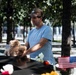 A man wearing a light blue t-shirt and brown baseball cap places a stuffed teddy bear on what appears to be the 9/11 Memorial in New York City. The dark granite memorial edge is adorned with flowers, small American flags, and other tributes. The man's downward gaze and deliberate gesture suggest a solemn, personal act of remembrance.