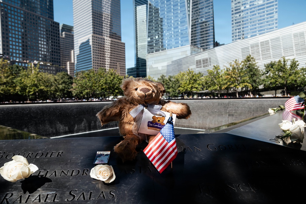 A memorial scene at the 9/11 Memorial in New York City, showing tributes placed on the engraved bronze parapet surrounding one of the reflecting pools. A brown teddy bear labeled 'The Comfort Cub' holds a small American flag, surrounded by white roses and other mementos. The Manhattan skyline, including One World Trade Center, is visible in the background.