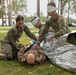 Three military personnel in US Army camouflage uniforms are conducting a combat casualty care training exercise outdoors. Two soldiers wearing simulated injury bandages around their heads and eyes are treating a prone soldier on the ground, using a mylar emergency blanket and what appears to be a tourniquet. The setting appears to be a military installation surrounded by pine trees.