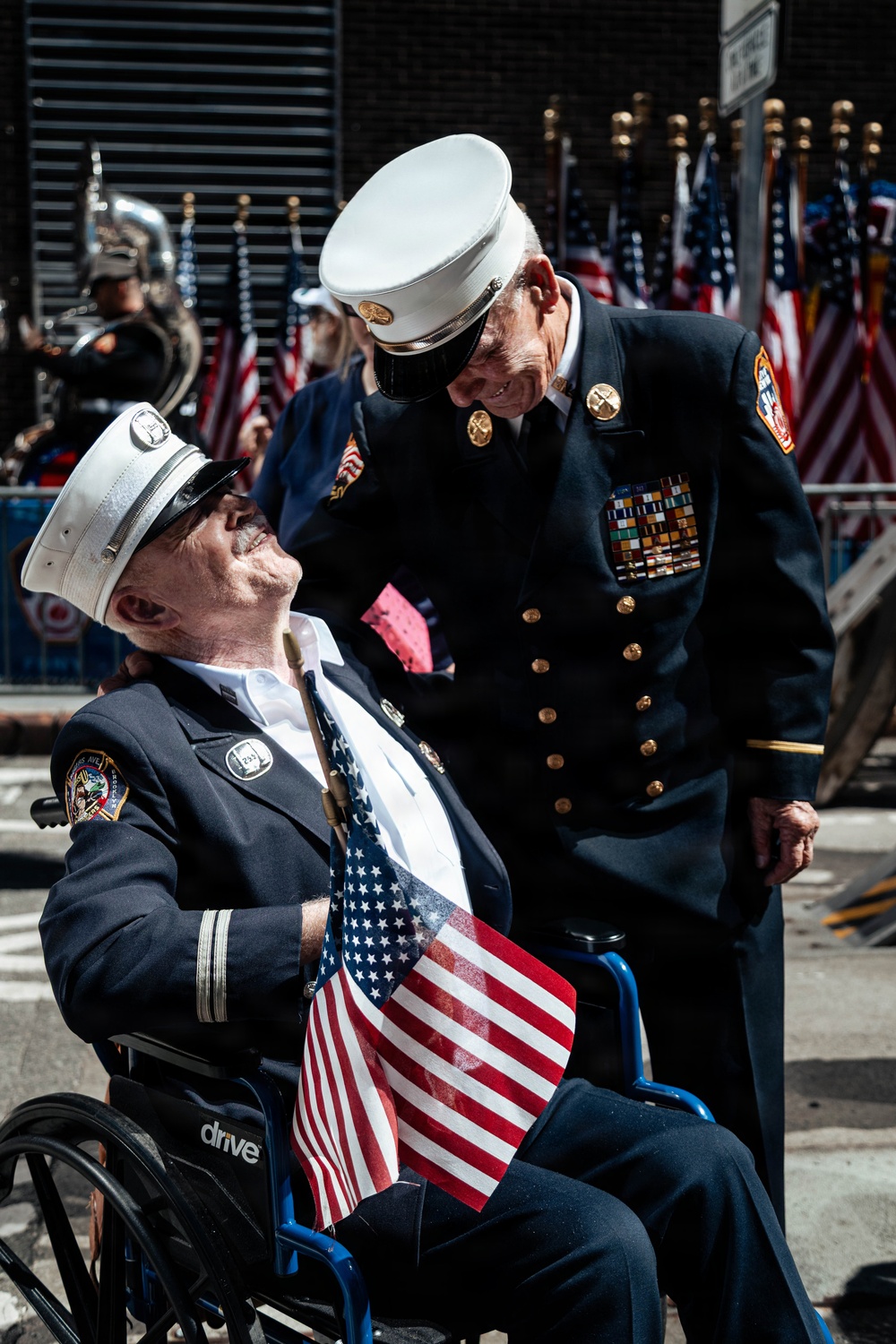 Two elderly men in formal fire department dress uniforms are sharing a moment at what appears to be a patriotic parade or ceremony. One man is seated in a 'Drive' brand wheelchair holding an American flag, while the other leans down toward him in conversation. Multiple American flags and a marching band are visible in the background.
