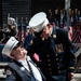 Two elderly men in formal fire department dress uniforms are sharing a moment at what appears to be a patriotic parade or ceremony. One man is seated in a 'Drive' brand wheelchair holding an American flag, while the other leans down toward him in conversation. Multiple American flags and a marching band are visible in the background.