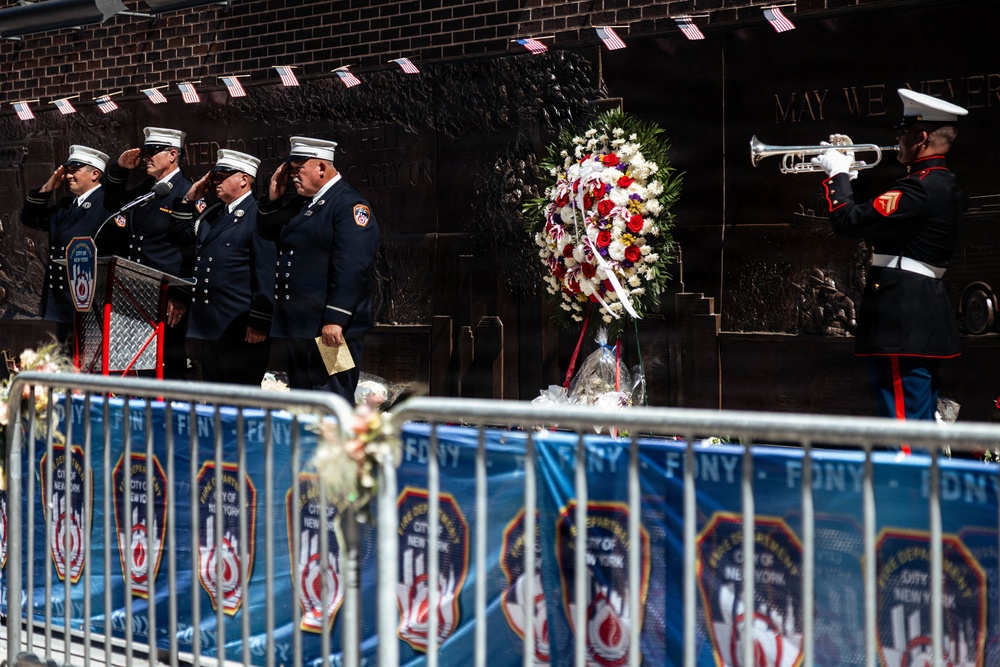 A solemn memorial ceremony is taking place at what appears to be an FDNY (Fire Department of New York) memorial wall. Four firefighters in dress uniforms are saluting while a Marine in dress blues plays a bugle, with a large floral wreath of red, white, and blue flowers placed before a bronze memorial wall. Small American flags are visible along the top of the wall, and FDNY-branded barriers line the foreground.