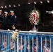 A solemn memorial ceremony is taking place at what appears to be an FDNY (Fire Department of New York) memorial wall. Four firefighters in dress uniforms are saluting while a Marine in dress blues plays a bugle, with a large floral wreath of red, white, and blue flowers placed before a bronze memorial wall. Small American flags are visible along the top of the wall, and FDNY-branded barriers line the foreground.