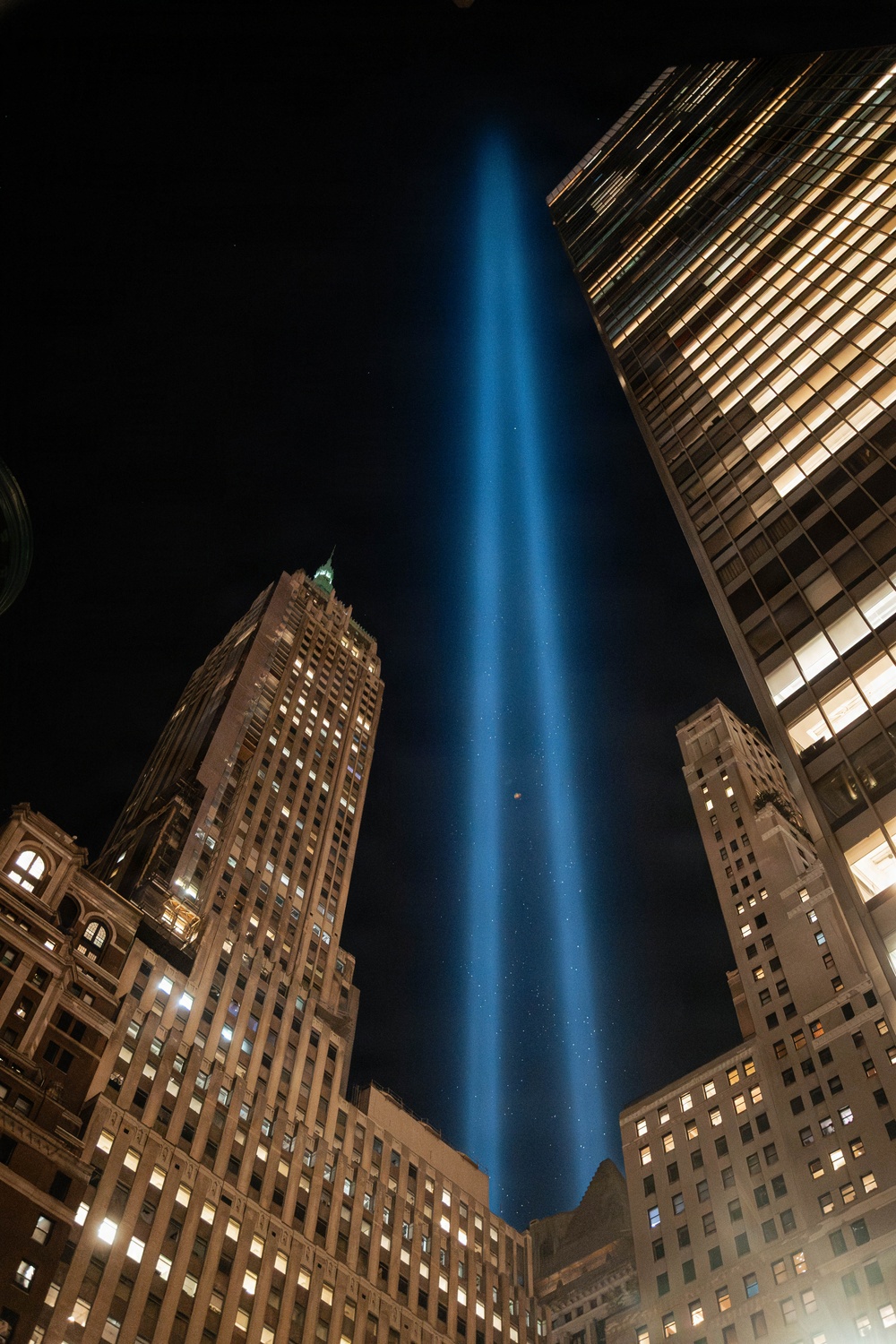 A nighttime urban photograph showing a powerful blue light beam projecting vertically into a dark sky, surrounded by illuminated skyscrapers. The composition is taken from a low angle looking upward, emphasizing the height and intensity of the beam against the city skyline. The scene is consistent with the 'Tribute in Light' memorial installation associated with the September 11 memorial in New York City.