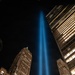 A nighttime urban photograph showing a powerful blue light beam projecting vertically into a dark sky, surrounded by illuminated skyscrapers. The composition is taken from a low angle looking upward, emphasizing the height and intensity of the beam against the city skyline. The scene is consistent with the 'Tribute in Light' memorial installation associated with the September 11 memorial in New York City.
