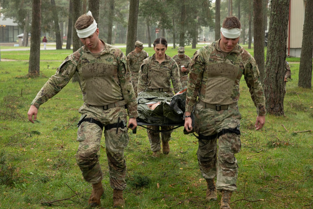 A group of military personnel in camouflage uniforms are conducting a field medical training exercise in a wooded area. Two soldiers wearing white head bandages are carrying a casualty on a litter/stretcher, while additional soldiers follow behind. The personnel appear wet and muddy, suggesting the exercise is being conducted in rainy, challenging conditions.