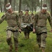 A group of military personnel in camouflage uniforms are conducting a field medical training exercise in a wooded area. Two soldiers wearing white head bandages are carrying a casualty on a litter/stretcher, while additional soldiers follow behind. The personnel appear wet and muddy, suggesting the exercise is being conducted in rainy, challenging conditions.