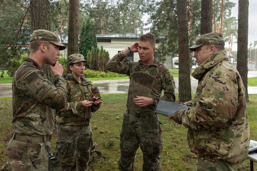 Four military personnel in OCP (Operational Camouflage Pattern) uniforms are engaged in a discussion outdoors near a wooded area. Three appear to be junior soldiers while one, holding a clipboard or tablet, appears to be a senior non-commissioned officer or officer conducting a briefing or after-action review. The setting appears to be a military installation in a European environment based on the architecture visible in the background.