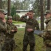 Four military personnel in OCP (Operational Camouflage Pattern) uniforms are engaged in a discussion outdoors near a wooded area. Three appear to be junior soldiers while one, holding a clipboard or tablet, appears to be a senior non-commissioned officer or officer conducting a briefing or after-action review. The setting appears to be a military installation in a European environment based on the architecture visible in the background.