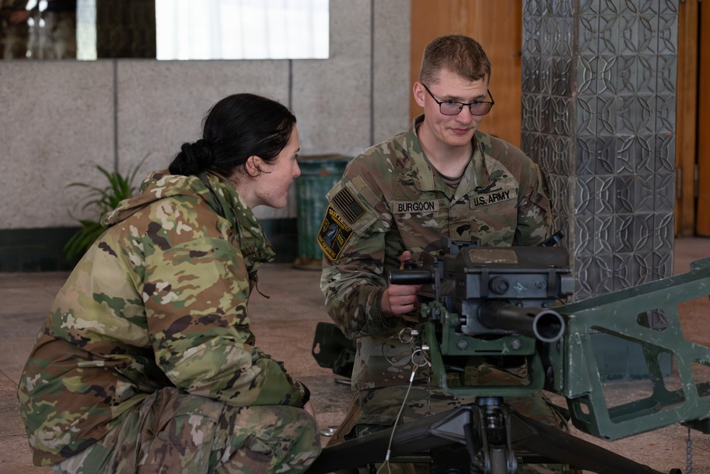 Two U.S. Army soldiers in OCP (Operational Camouflage Pattern) uniforms are engaged in what appears to be weapons system training or maintenance. The soldier on the right, identified by name tape as 'BURGOON,' is demonstrating or explaining the operation of a mounted weapon system to the other soldier. The setting appears to be an indoor or covered outdoor facility.