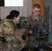 Two U.S. Army soldiers in OCP (Operational Camouflage Pattern) uniforms are engaged in what appears to be weapons system training or maintenance. The soldier on the right, identified by name tape as 'BURGOON,' is demonstrating or explaining the operation of a mounted weapon system to the other soldier. The setting appears to be an indoor or covered outdoor facility.