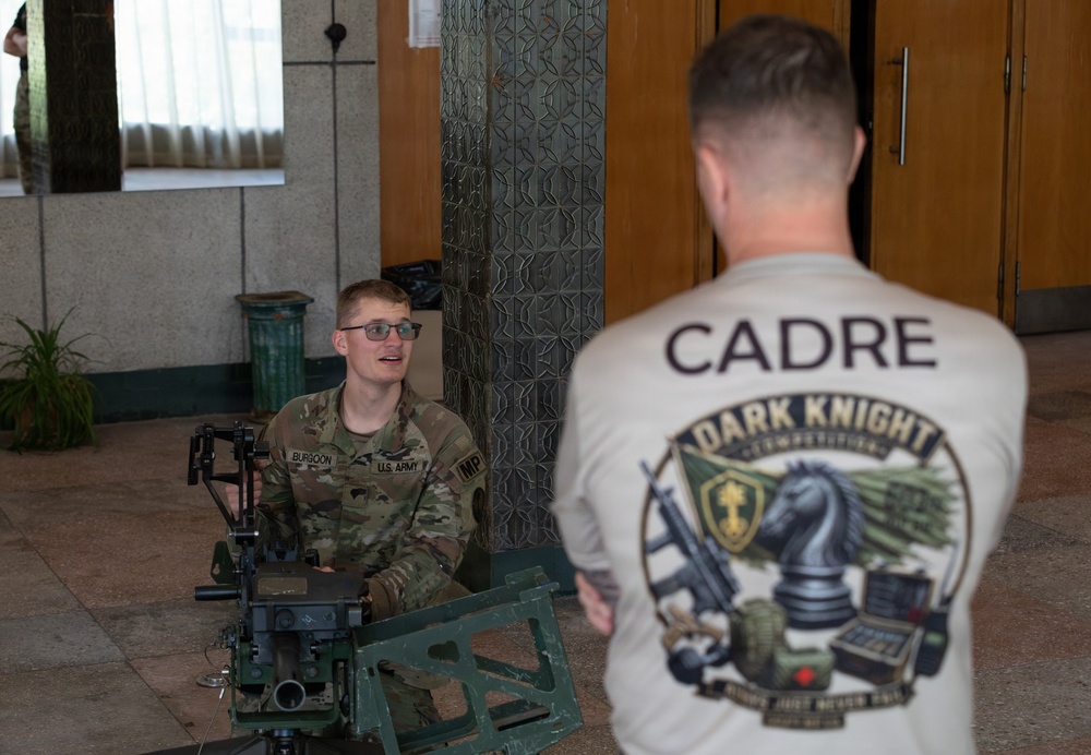 A U.S. Army Military Police soldier named Burgoon is seated on the floor working with a mounted weapon system, likely during a skills competition or evaluation. A cadre member wearing a 'Dark Knight Competition' shirt observes or evaluates the soldier. The setting appears to be an indoor facility with tiled floors and ornate doors.