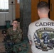 A U.S. Army Military Police soldier named Burgoon is seated on the floor working with a mounted weapon system, likely during a skills competition or evaluation. A cadre member wearing a 'Dark Knight Competition' shirt observes or evaluates the soldier. The setting appears to be an indoor facility with tiled floors and ornate doors.
