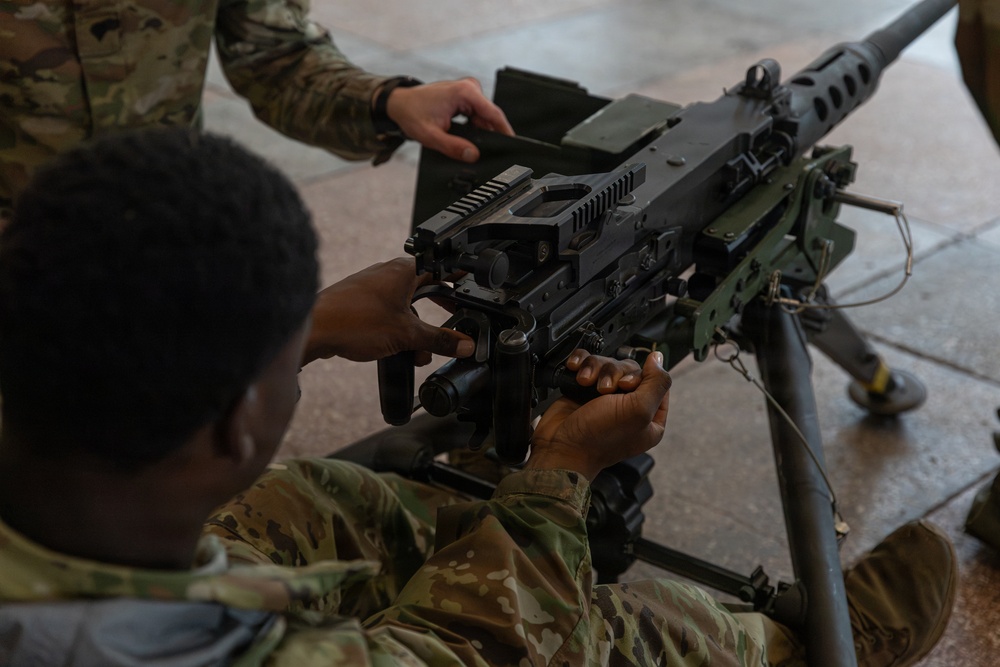 Two military personnel in OCP (Operational Camouflage Pattern) uniforms are handling a crew-served machine gun mounted on a tripod. The image appears to depict a training or familiarization exercise, with one soldier positioned behind the weapon and another assisting from above. The weapon is mounted on a standard military M3 tripod.