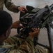 Two military personnel in OCP (Operational Camouflage Pattern) uniforms are handling a crew-served machine gun mounted on a tripod. The image appears to depict a training or familiarization exercise, with one soldier positioned behind the weapon and another assisting from above. The weapon is mounted on a standard military M3 tripod.