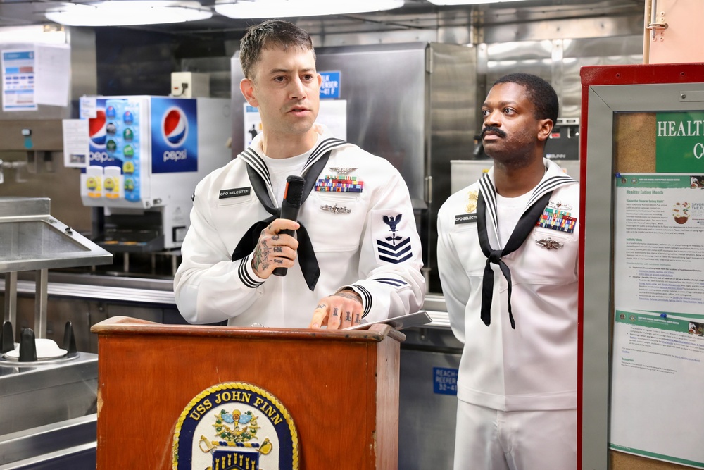 Two U.S. Navy sailors in dress white uniforms stand at a wooden podium bearing the USS John Finn ship's seal during what appears to be an official ceremony or briefing. The sailor at the podium is holding a microphone and wearing a 'CPO SELECTEE' name tag along with multiple service ribbons. The setting appears to be a shipboard galley or dining facility.