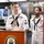 Two U.S. Navy sailors in dress white uniforms stand at a wooden podium bearing the USS John Finn ship's seal during what appears to be an official ceremony or briefing. The sailor at the podium is holding a microphone and wearing a 'CPO SELECTEE' name tag along with multiple service ribbons. The setting appears to be a shipboard galley or dining facility.