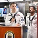 Two U.S. Navy sailors in dress white uniforms stand at a wooden podium bearing the USS John Finn ship's seal during what appears to be an official ceremony or briefing. The sailor at the podium is holding a microphone and wearing a 'CPO SELECTEE' name tag along with multiple service ribbons. The setting appears to be a shipboard galley or dining facility.