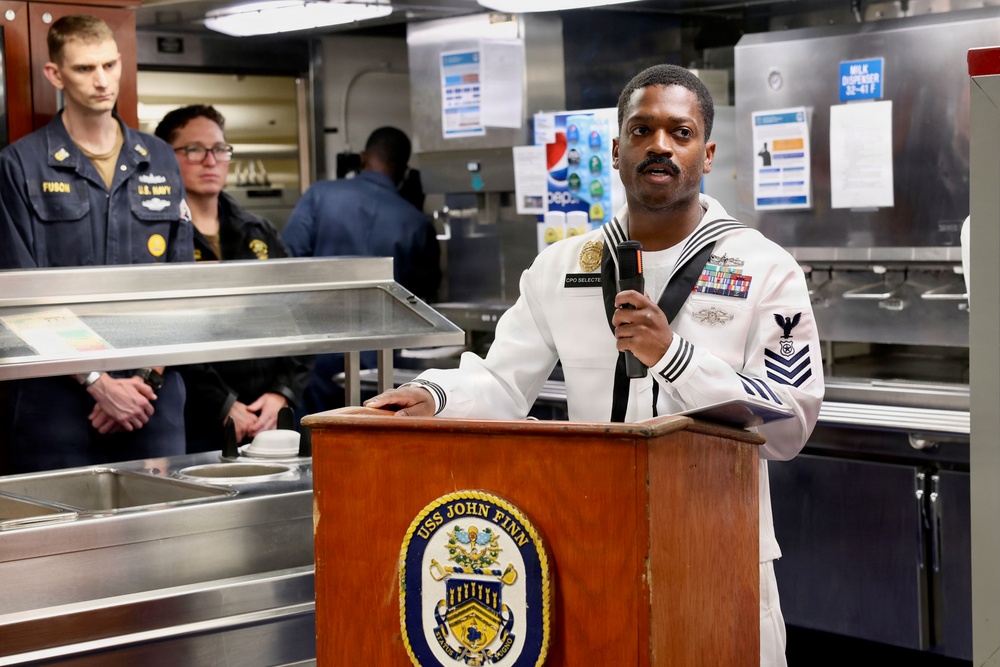 A U.S. Navy sailor in dress white uniform speaks at a podium bearing the USS John Finn ship's crest inside what appears to be the ship's mess hall or galley. Two other Navy personnel in working khaki uniforms observe from the background. The setting suggests an official ceremony or formal address aboard the destroyer USS John Finn.