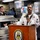 A U.S. Navy sailor in dress white uniform speaks at a podium bearing the USS John Finn ship's crest inside what appears to be the ship's mess hall or galley. Two other Navy personnel in working khaki uniforms observe from the background. The setting suggests an official ceremony or formal address aboard the destroyer USS John Finn.