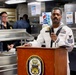 A U.S. Navy sailor in dress white uniform speaks at a podium bearing the USS John Finn ship's crest inside what appears to be the ship's mess hall or galley. Two other Navy personnel in working khaki uniforms observe from the background. The setting suggests an official ceremony or formal address aboard the destroyer USS John Finn.
