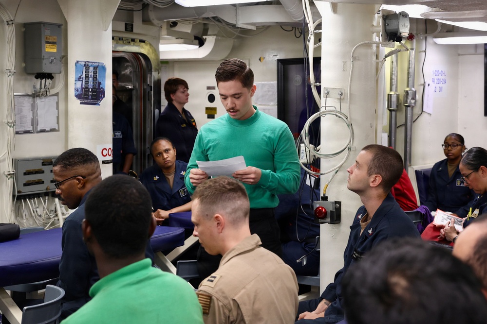 A group of military personnel, predominantly in navy blue uniforms, are gathered in what appears to be a briefing or meeting room aboard a naval vessel. A young man in a green sweater stands at the center, reading from a paper document, suggesting he is presenting or conducting a briefing. The setting features typical shipboard interior elements including pipes, conduits, and institutional lighting.
