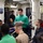A group of military personnel, predominantly in navy blue uniforms, are gathered in what appears to be a briefing or meeting room aboard a naval vessel. A young man in a green sweater stands at the center, reading from a paper document, suggesting he is presenting or conducting a briefing. The setting features typical shipboard interior elements including pipes, conduits, and institutional lighting.