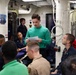 A group of military personnel, predominantly in navy blue uniforms, are gathered in what appears to be a briefing or meeting room aboard a naval vessel. A young man in a green sweater stands at the center, reading from a paper document, suggesting he is presenting or conducting a briefing. The setting features typical shipboard interior elements including pipes, conduits, and institutional lighting.