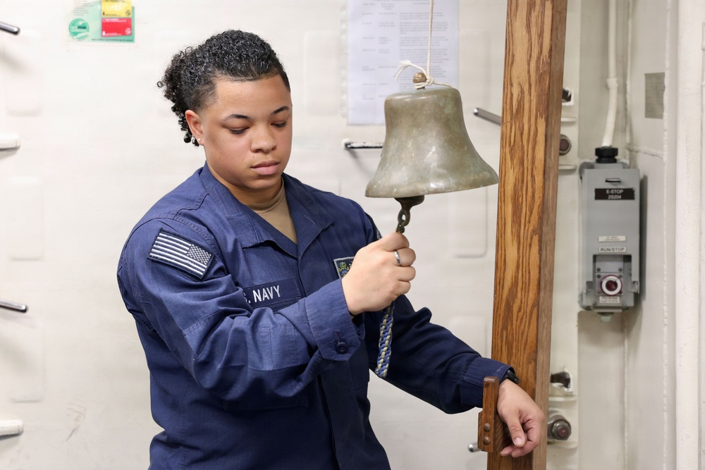 A U.S. Navy sailor in blue working uniform is ringing a ship's bell aboard a naval vessel. The sailor is holding the bell's clapper rope and appears to be performing an official duty, likely marking the time or signaling a watch change. The setting appears to be an interior compartment of a naval ship.