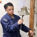 A U.S. Navy sailor in blue working uniform is ringing a ship's bell aboard a naval vessel. The sailor is holding the bell's clapper rope and appears to be performing an official duty, likely marking the time or signaling a watch change. The setting appears to be an interior compartment of a naval ship.