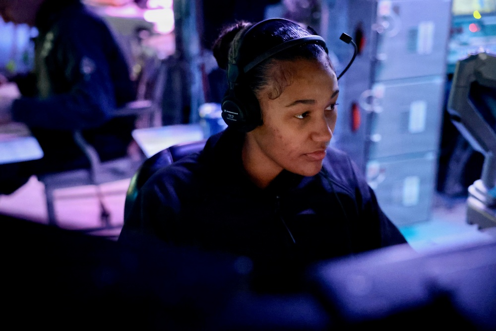 A person wearing a headset with a boom microphone is seated at a workstation in a dimly lit, blue-toned operational environment. They appear focused on a monitor in front of them, suggesting a communications or monitoring role. The setting resembles a military operations center, broadcast booth, or similar command-and-control environment.