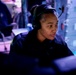 A person wearing a headset with a boom microphone is seated at a workstation in a dimly lit, blue-toned operational environment. They appear focused on a monitor in front of them, suggesting a communications or monitoring role. The setting resembles a military operations center, broadcast booth, or similar command-and-control environment.