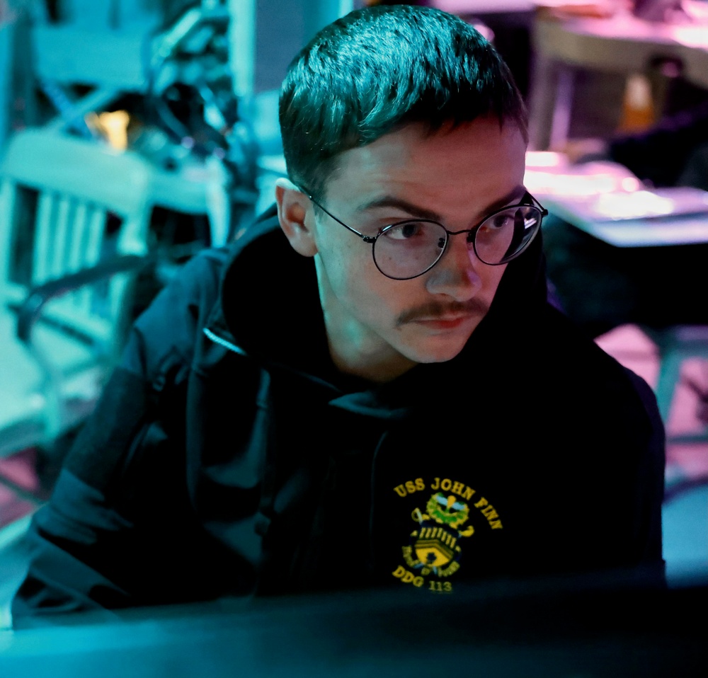 A young person with a mustache and round wire-framed glasses is focused intently on a screen, illuminated by blue-green and pink ambient lighting. They are wearing a black hoodie bearing the text 'USS JOHN FINN DDG 113', indicating a U.S. Navy destroyer affiliation. The environment appears to be a dimly lit operational or workstation setting.