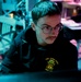 A young person with a mustache and round wire-framed glasses is focused intently on a screen, illuminated by blue-green and pink ambient lighting. They are wearing a black hoodie bearing the text 'USS JOHN FINN DDG 113', indicating a U.S. Navy destroyer affiliation. The environment appears to be a dimly lit operational or workstation setting.